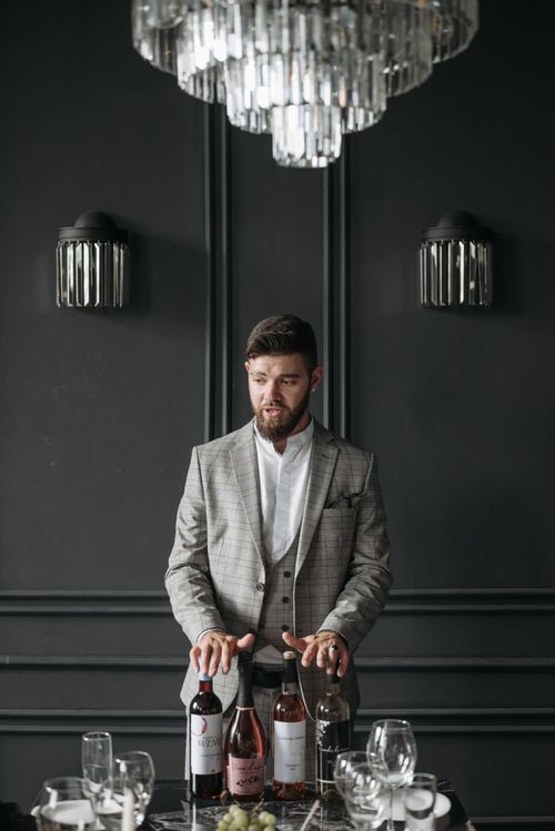 Free A man in a suit stands indoors with wine bottles, reflecting an elegant dinner arrangement. Stock Photo