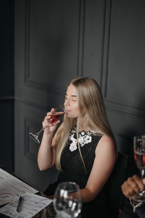 Blonde-Haired Woman in Black Dress Sitting while Drinking a Red Wine
