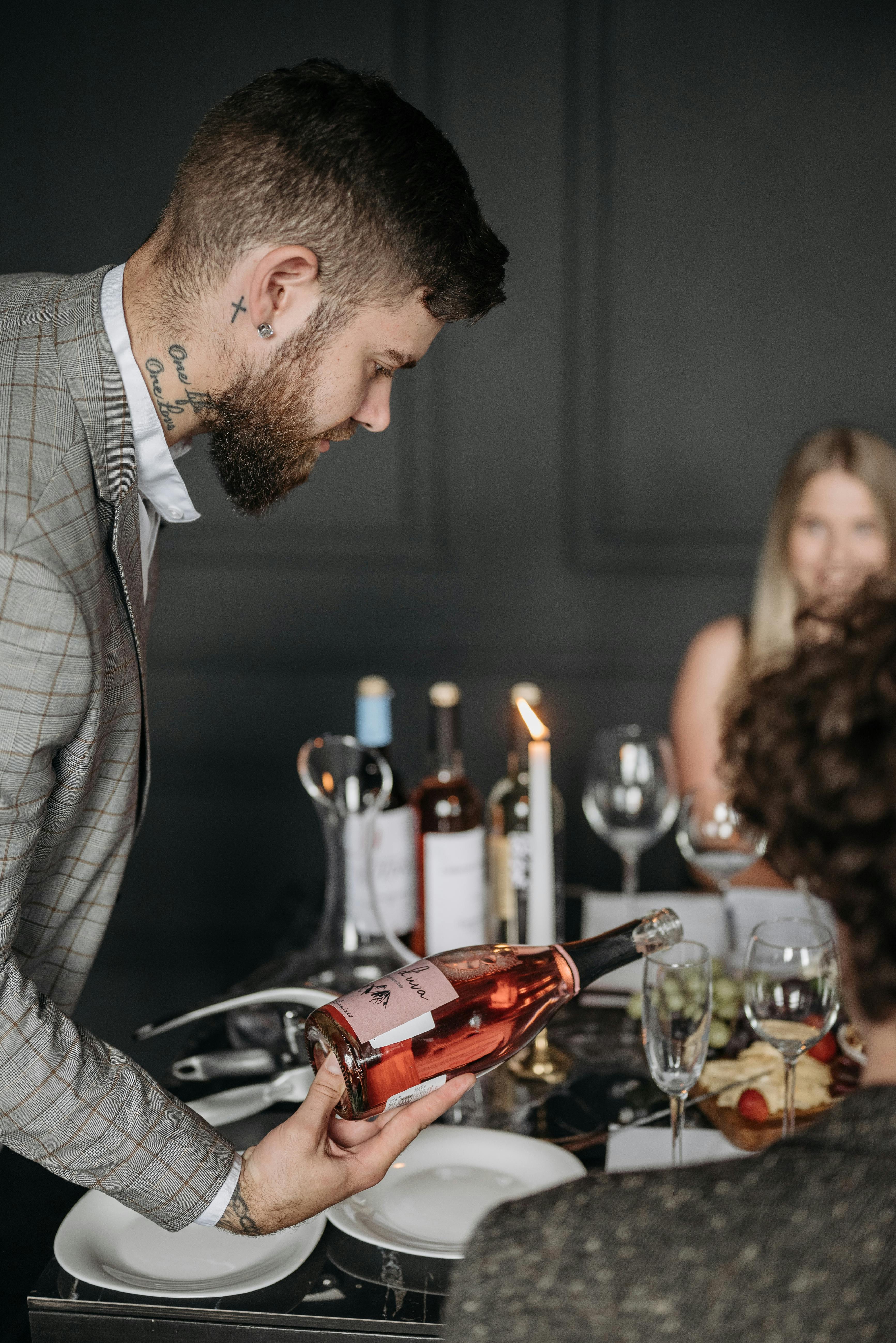 Caucasian man with tattoos serves wine at an elegant indoor dinner gathering.