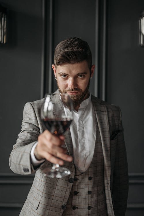 A Man in Gray Plaid Suit Holding a Goblet Glass with Red Wine while Looking at the Camera