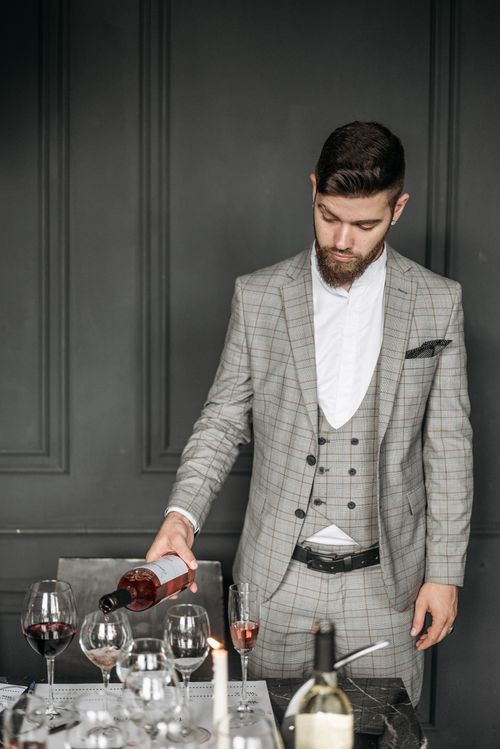 Free Bearded man in a plaid suit pouring red wine at an elegant indoor event setting. Stock Photo