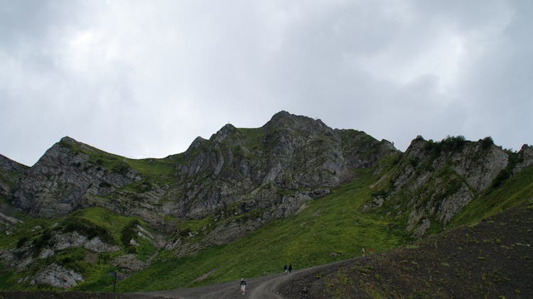 Photo Of People Walking On A Pathway Against The Background Of A Mountain Partially Covered With Grass