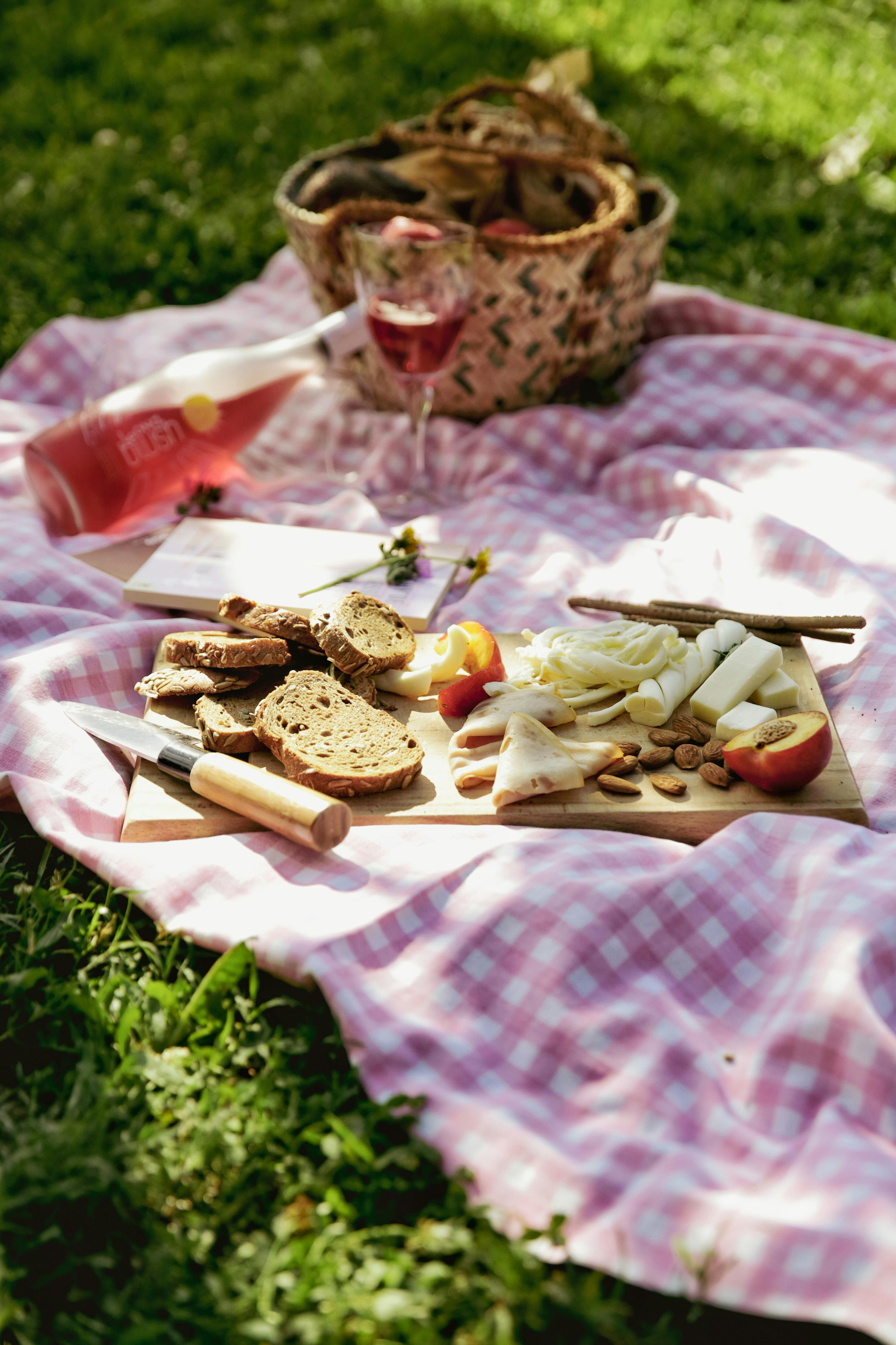 A delightful picnic setup on a pink checkered blanket with bread, cheese, and wine in a sunny park.