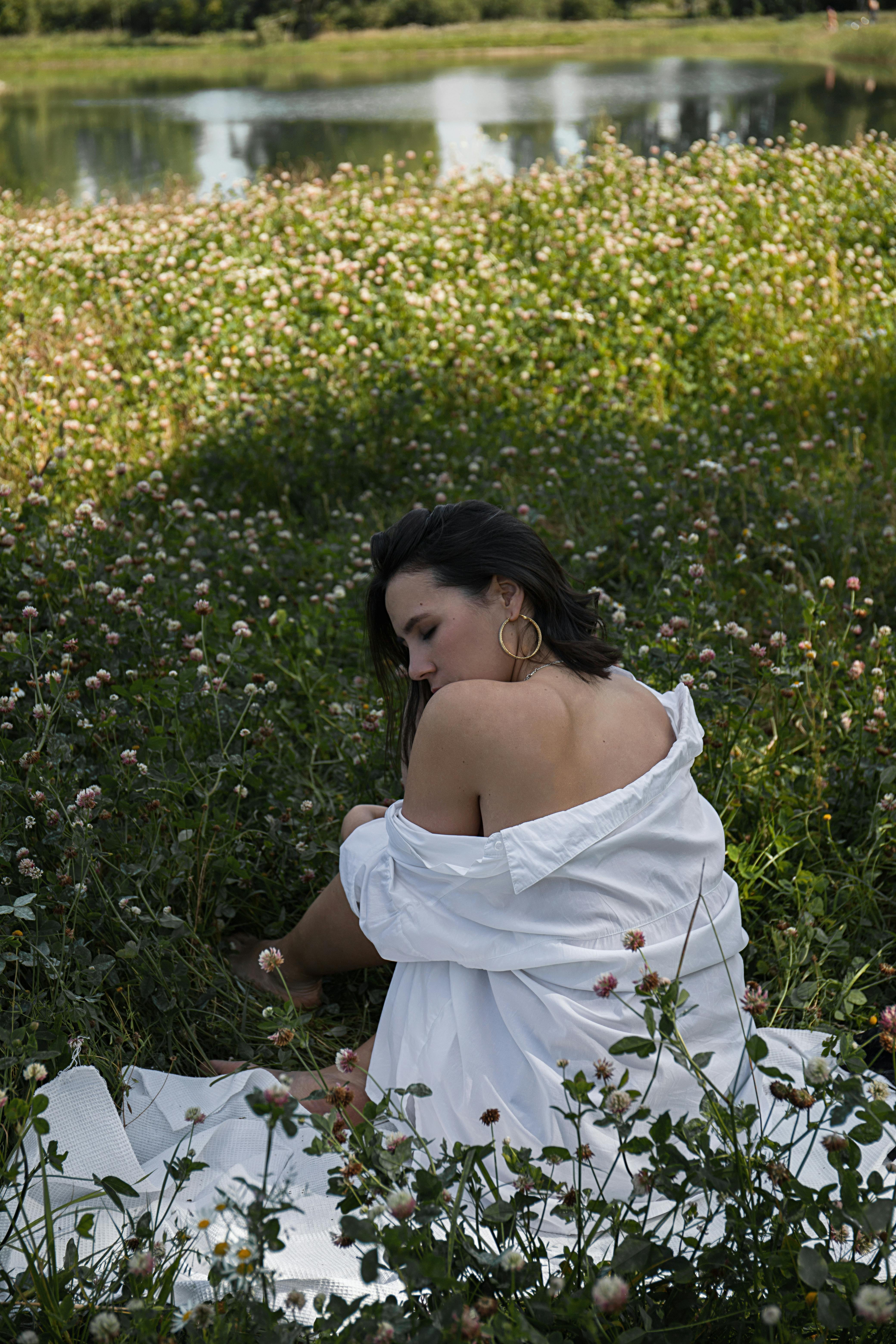 A woman in a white dress sits in a field of clover by a tranquil lake in summer.