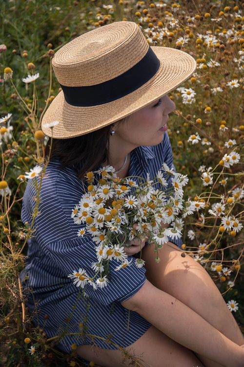 Free A woman wearing a straw hat sits in a field holding a bouquet of daisies, surrounded by blooming flowers. Stock Photo