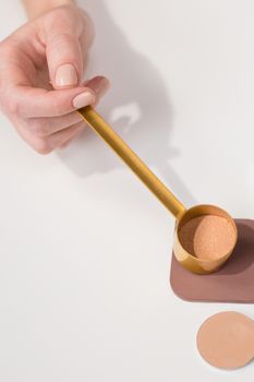 A hand holding a golden scoop of cosmetic powder on a white surface, minimalist style.