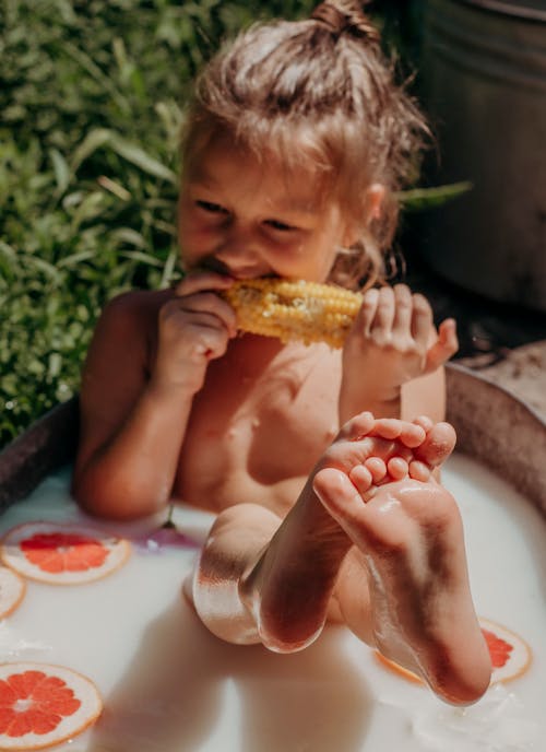 A Child Sitting on a Basin with Milk Bath while Eating Corn