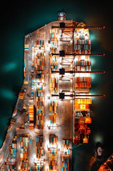 Dramatic aerial view of a busy shipping port with colorful containers and bright lights at night.