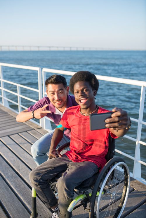 Free Two friends, one in a wheelchair, taking a selfie on a sunny boardwalk by the water. Stock Photo