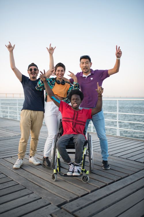 Free Group of diverse friends, including a person in a wheelchair, joyfully posing outdoors by the waterfront. Stock Photo