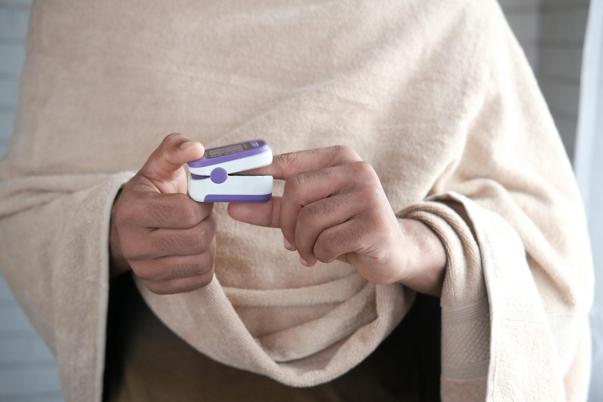 Adult hands measuring oxygen levels using a pulse oximeter, wrapped in a blanket indoors.