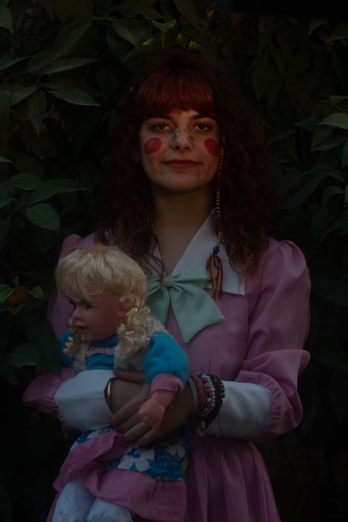 Free A woman with painted cheeks in a pink dress holding a doll amidst green foliage. Stock Photo