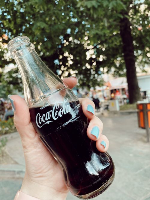 Free A hand with manicured nails holds a glass Coca-Cola bottle in an outdoor setting. Stock Photo