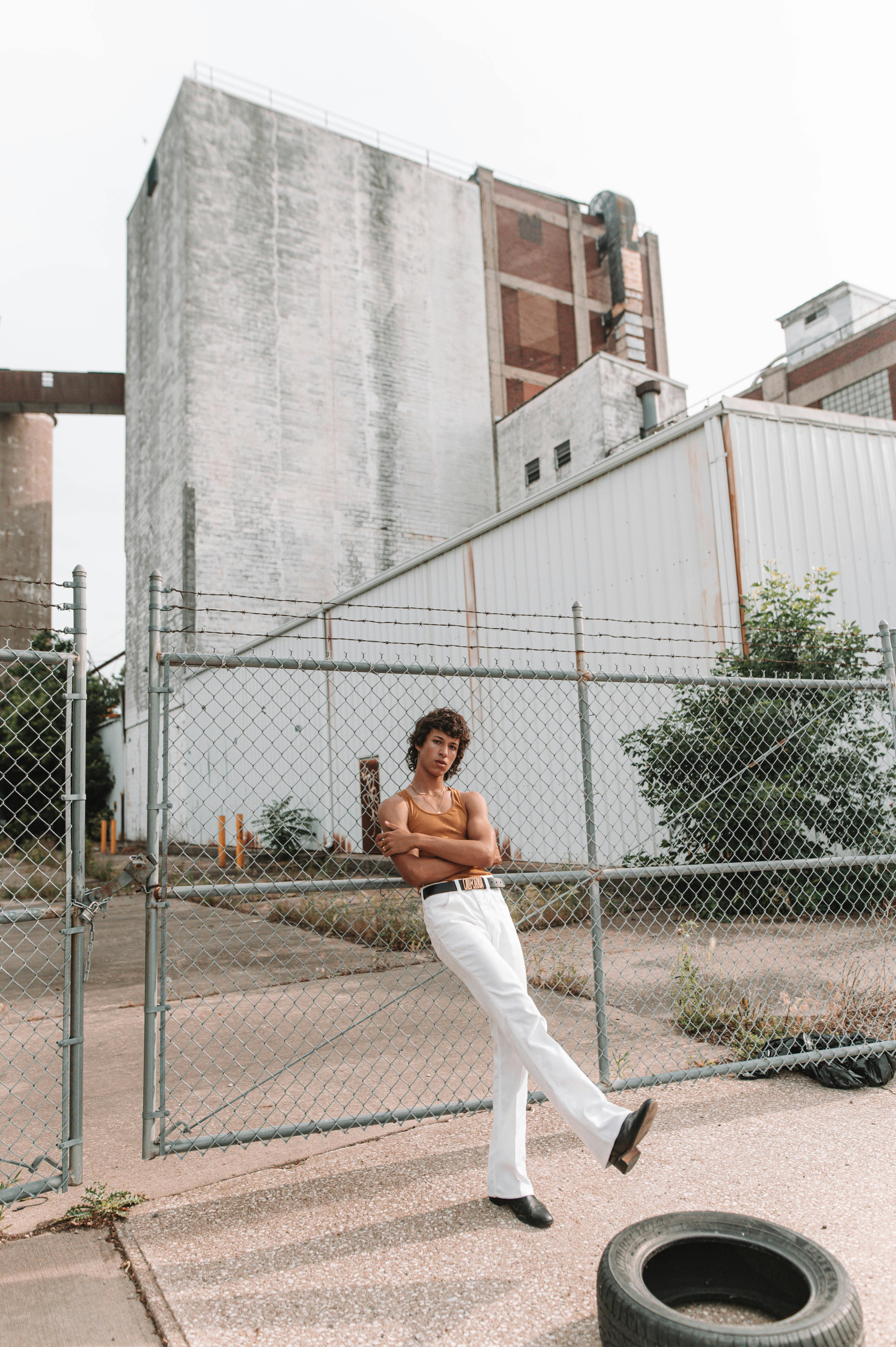 A man in white pants and black shoes poses confidently before an industrial backdrop.