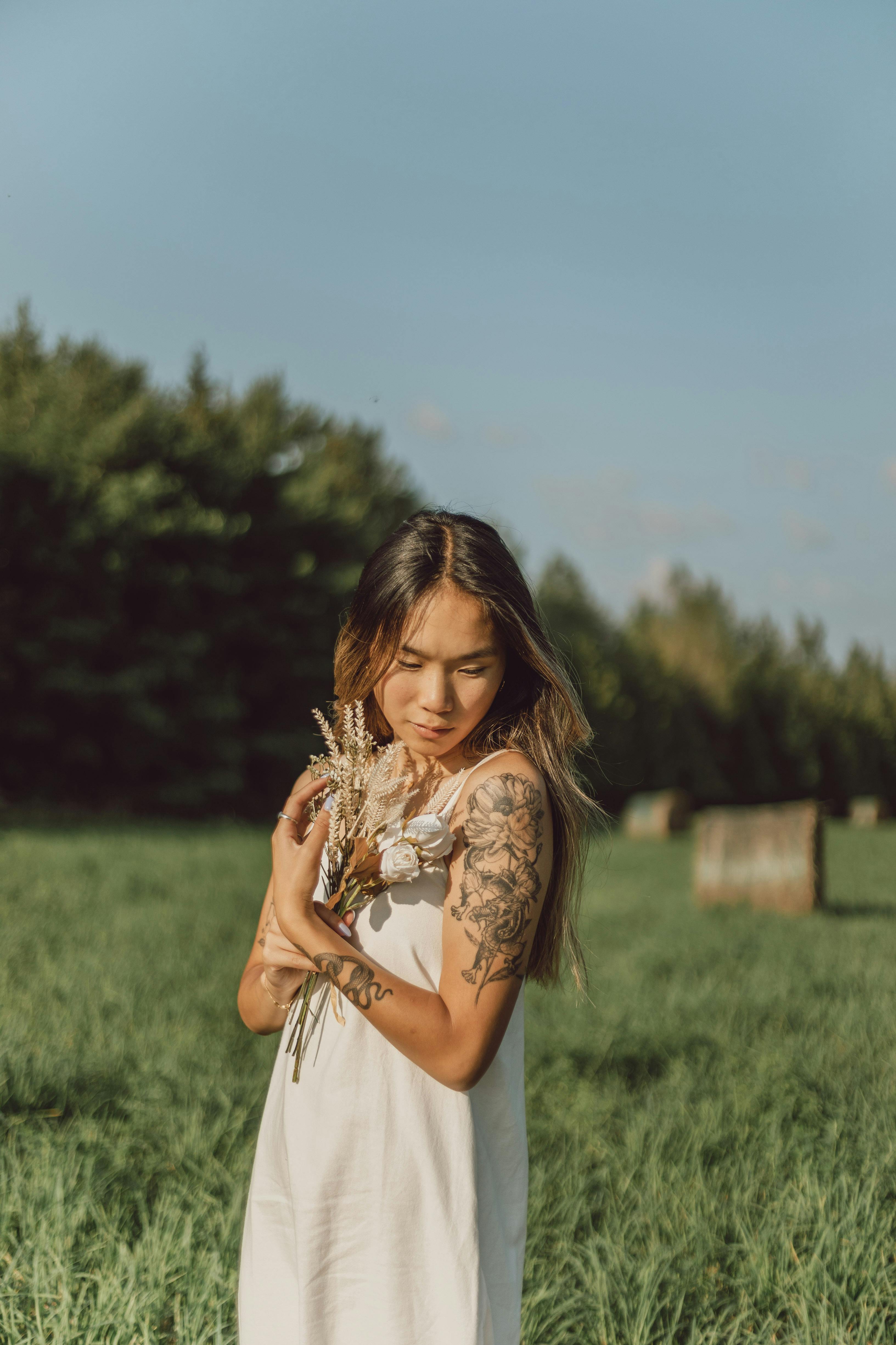 A serene portrait of a young woman in a white dress, standing in a rural field with wildflowers.