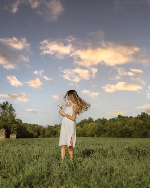 Woman in White Dress Holding a Flower While in a Grass Land