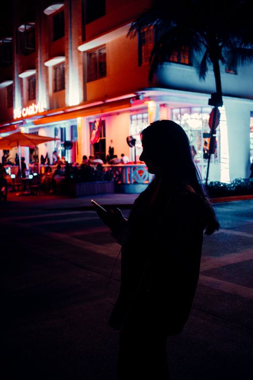 Free A woman silhouetted against a vibrant city street, using her phone at night. Stock Photo