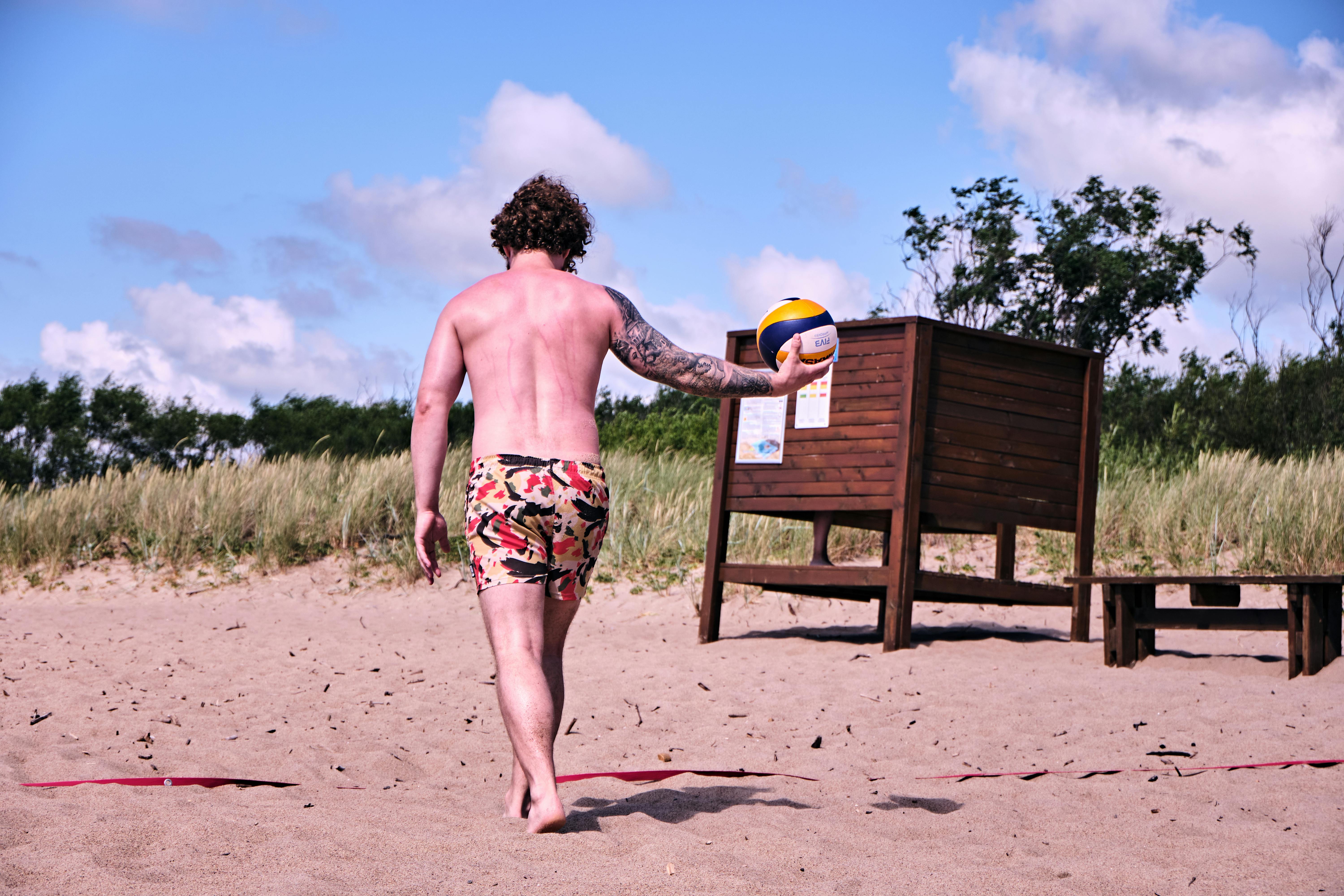 a shirtless man walking on the beach sand while holding a volleyball