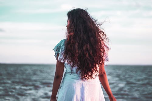 Back View of a Curly-Haired Woman in White Dress Standing in Front of Ocean