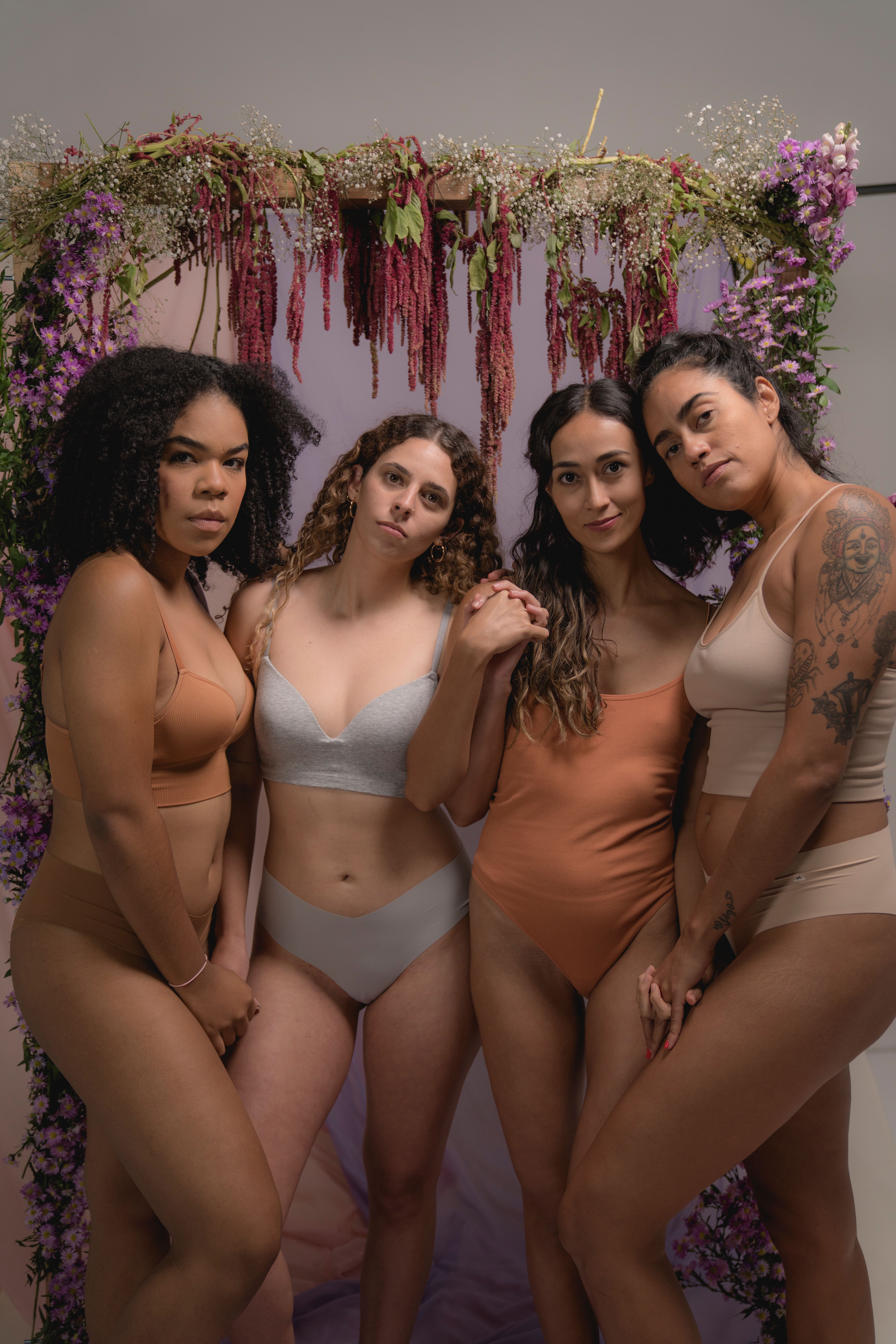 A group of diverse women in lingerie poses together before a floral archway.