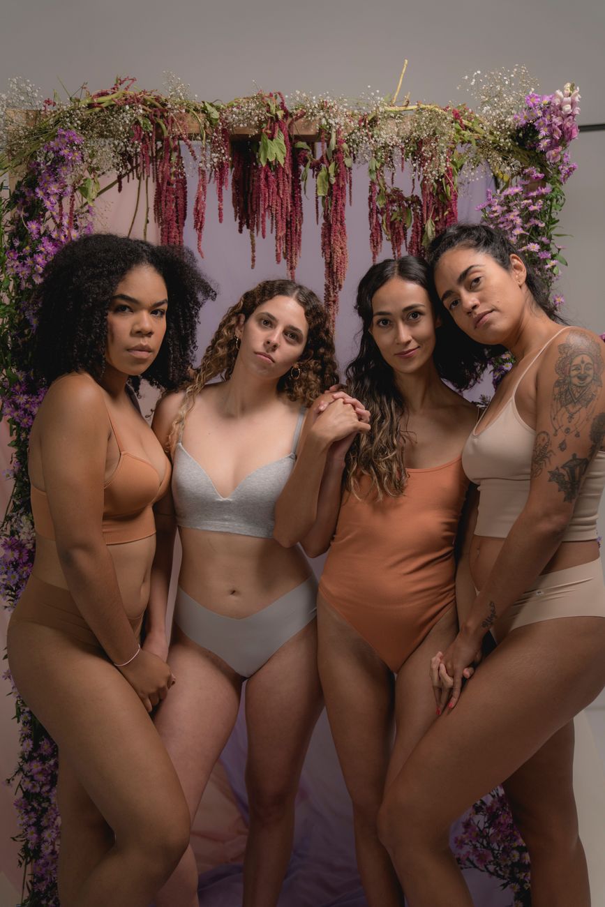 A group of diverse women in lingerie poses together before a floral archway.