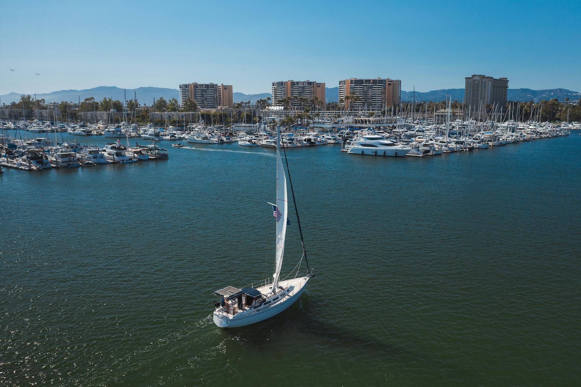 Aerial shot of a sailboat in Marina del Rey with urban skyline background.