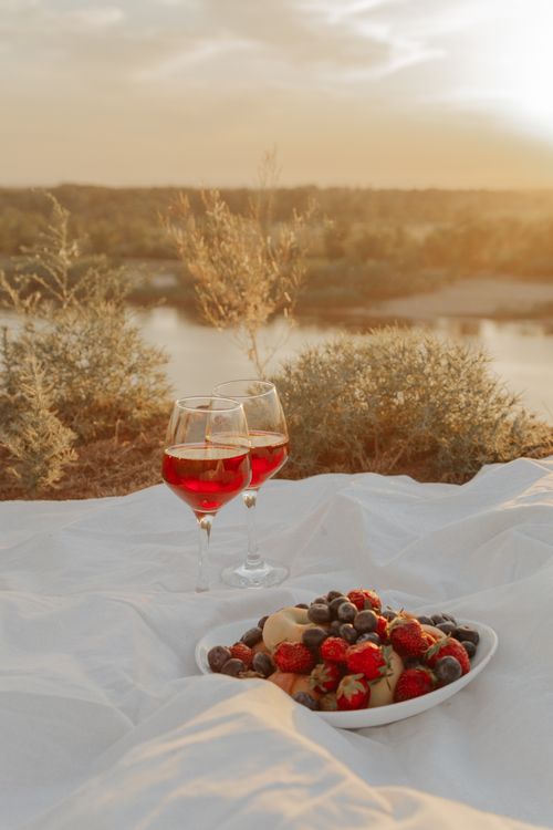 Free A serene lakeside picnic at sunset featuring a bowl of fresh fruits and glasses of wine on a blanket. Stock Photo