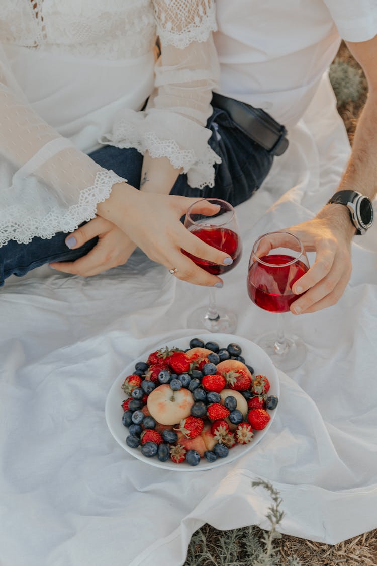 Couple Drinking Red Fruit Juice On Picnic And Various Fruits On A White Blanket