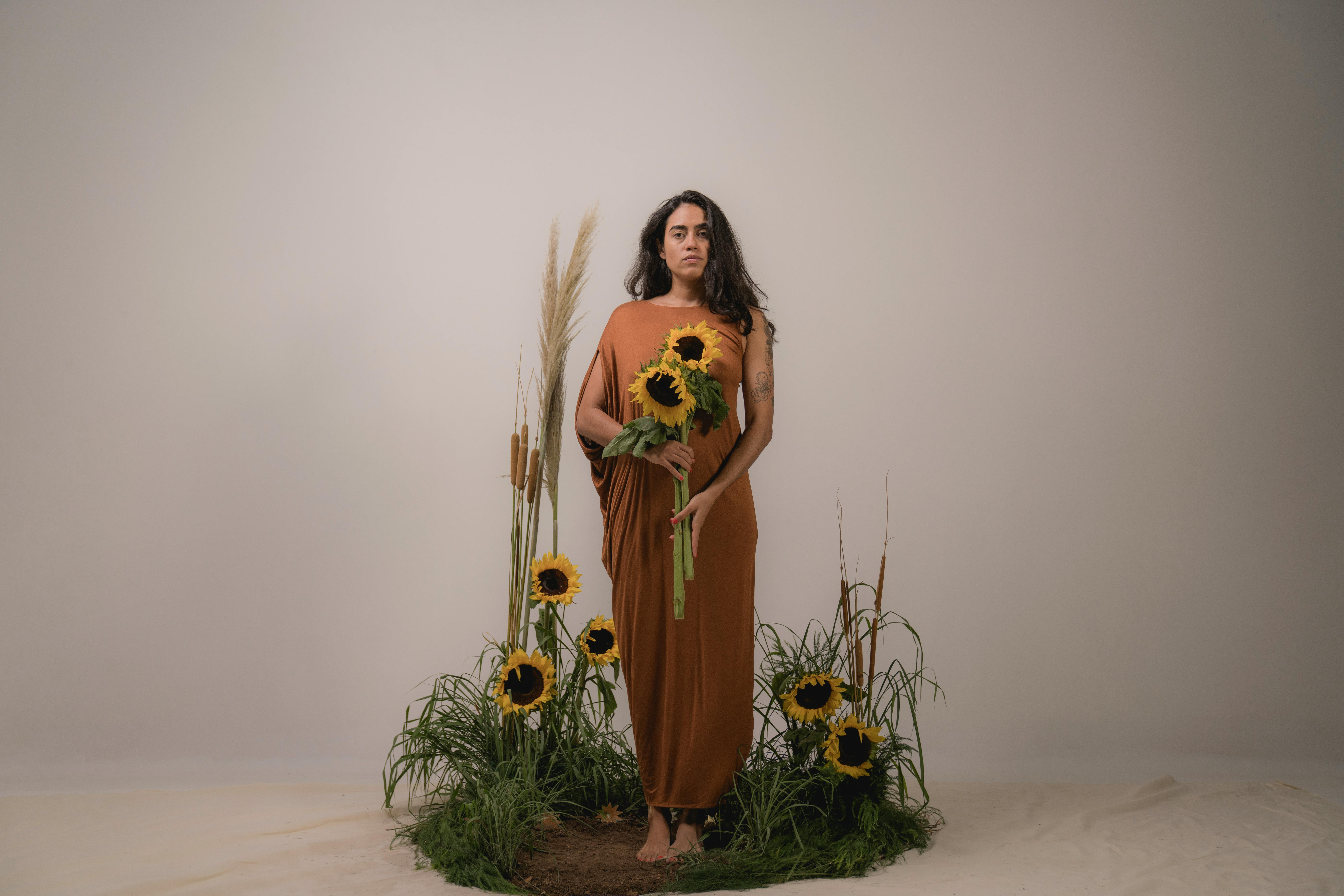 An elegant woman in a brown dress poses with sunflowers in a studio setting.