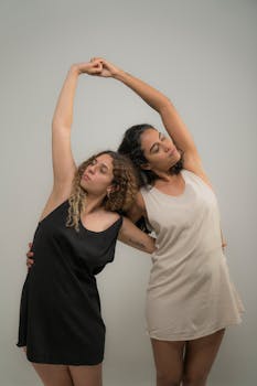 Two women with curly hair gracefully stretching in dresses against a plain background.