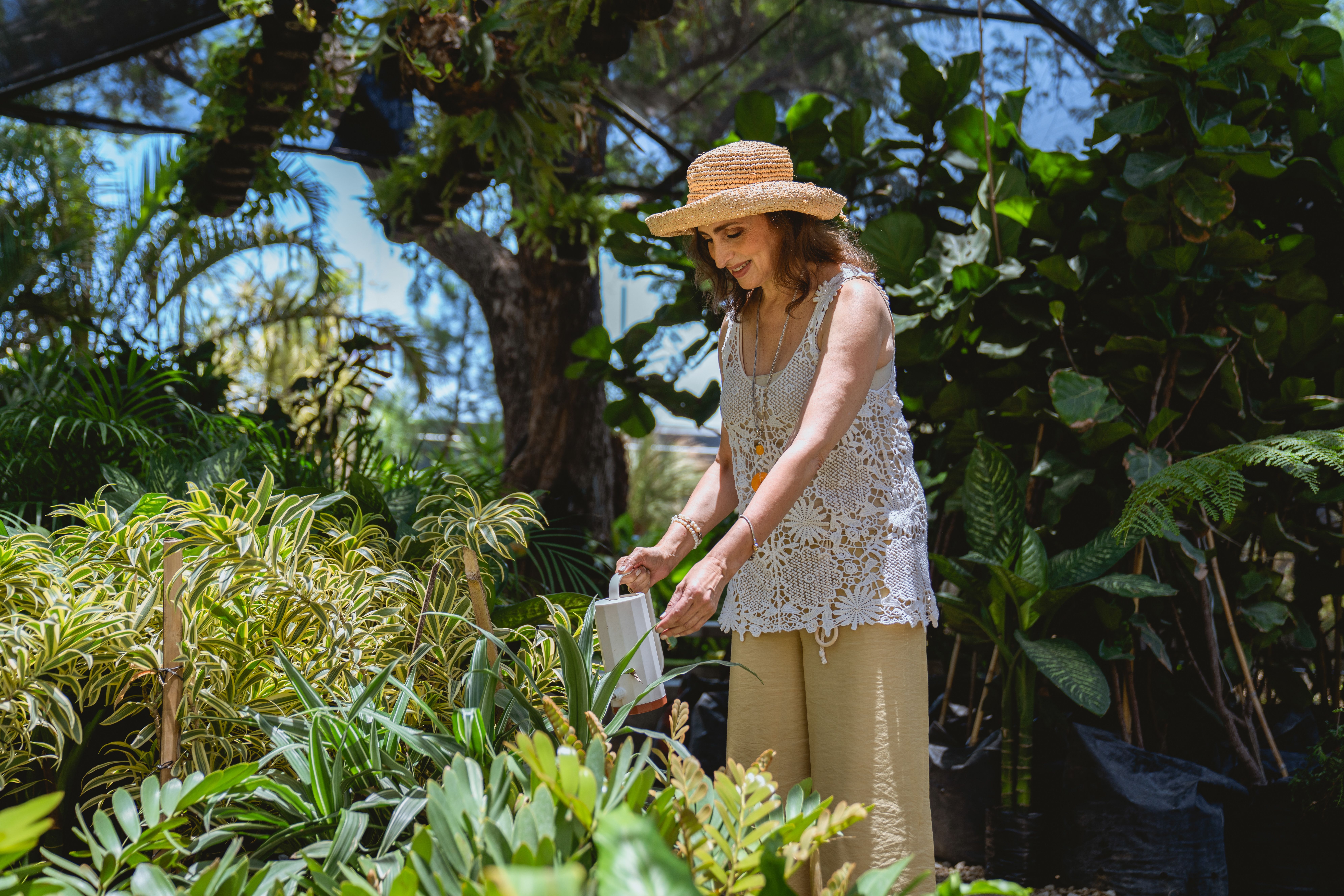 Elderly woman watering plants with a smile in a vibrant greenhouse setting.