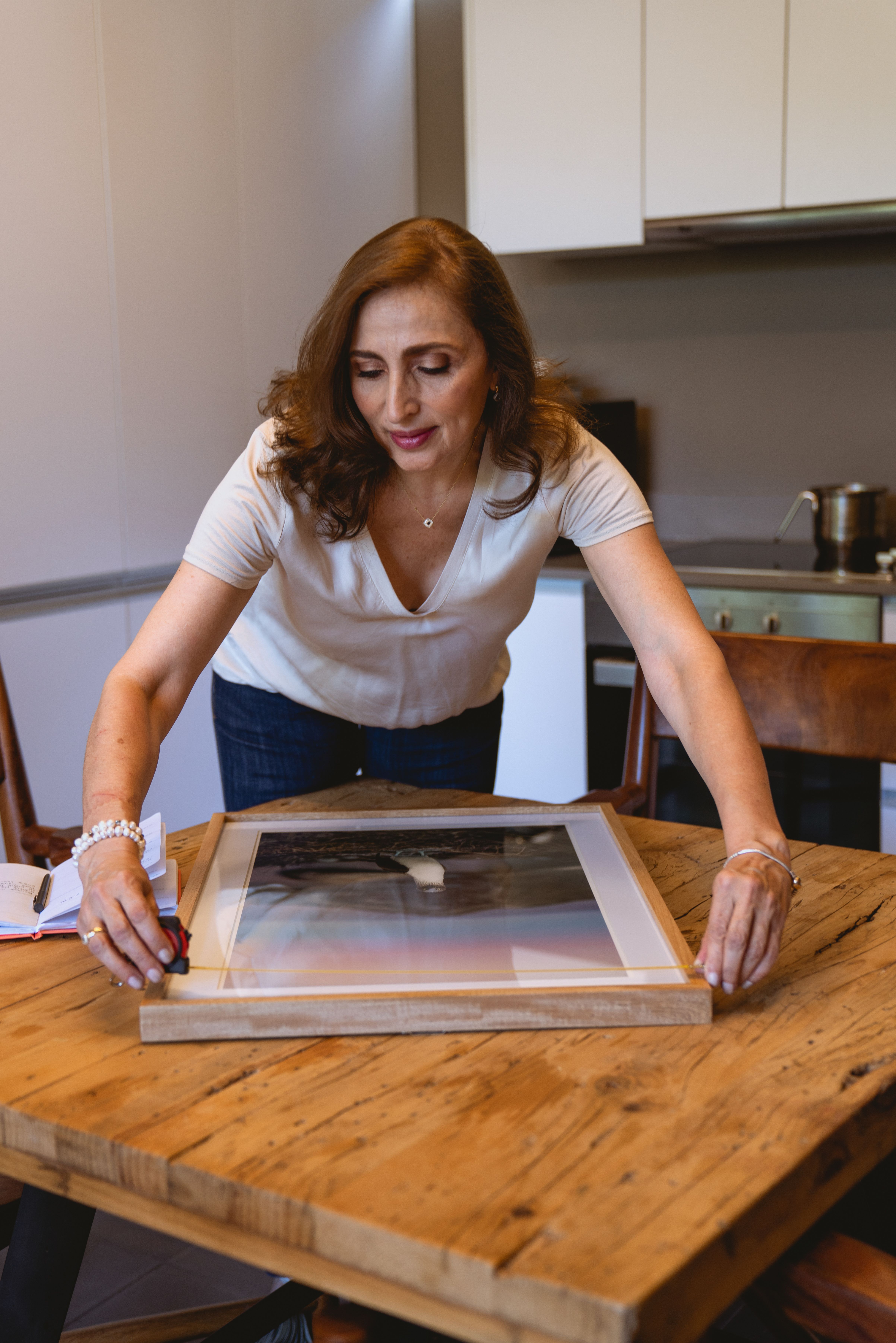 Woman in white shirt adjusting a frame on wooden table inside modern kitchen.