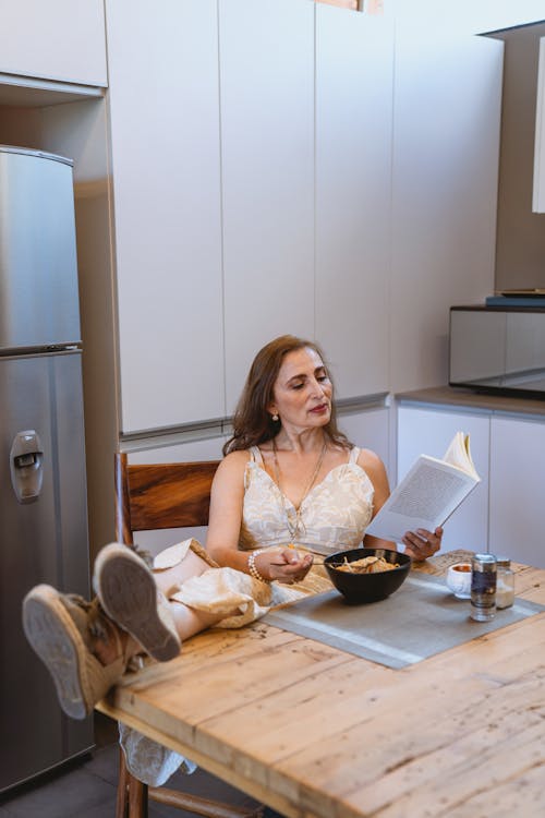 Free Woman Eating While Reading a Book With Feet on the Table Stock Photo