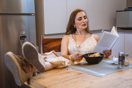 Free Woman in White Tank Top Eating While Reading a Book With Feet on the Table Stock Photo