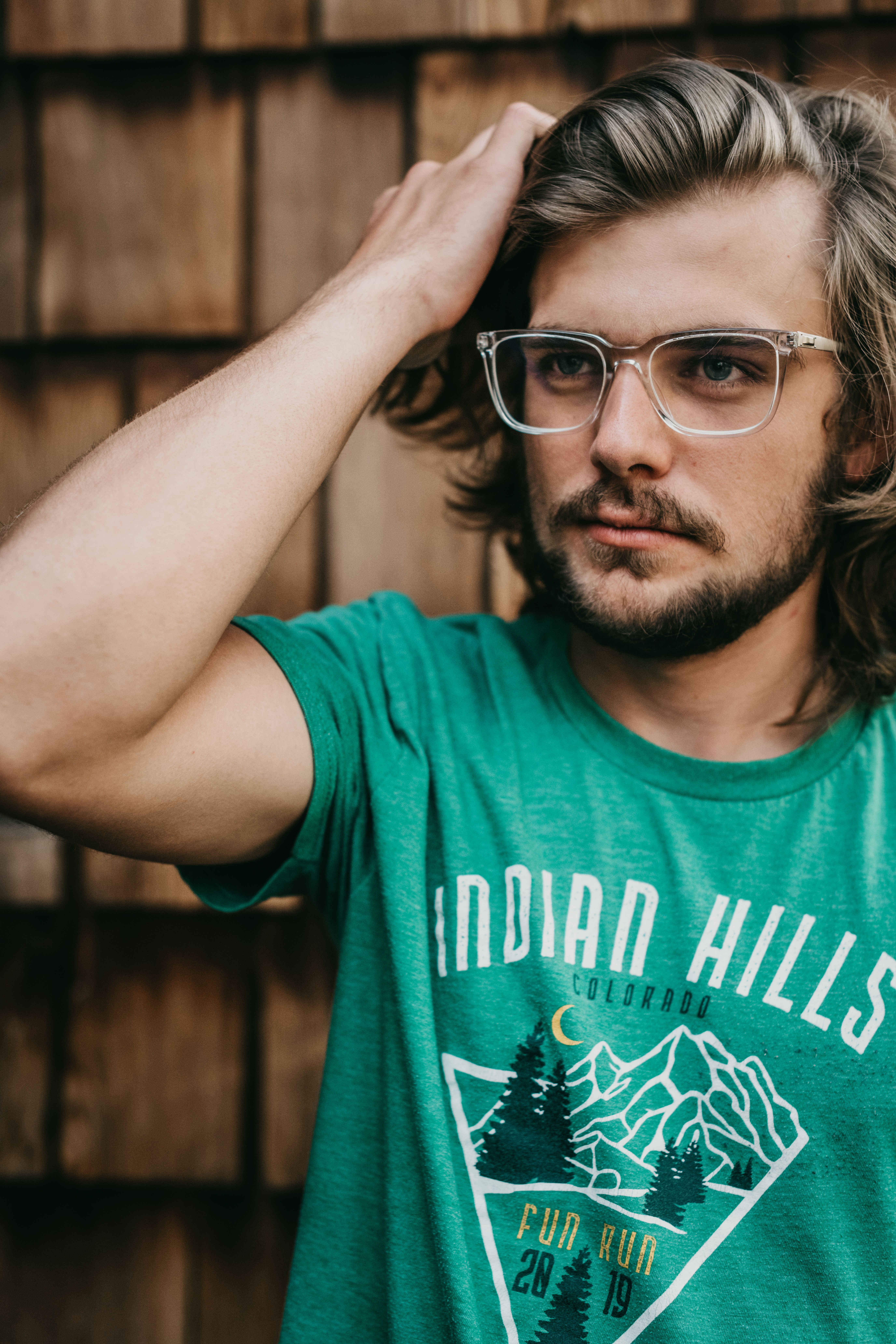 A casual portrait of a bearded man fixing his long hair while wearing glasses outdoors.