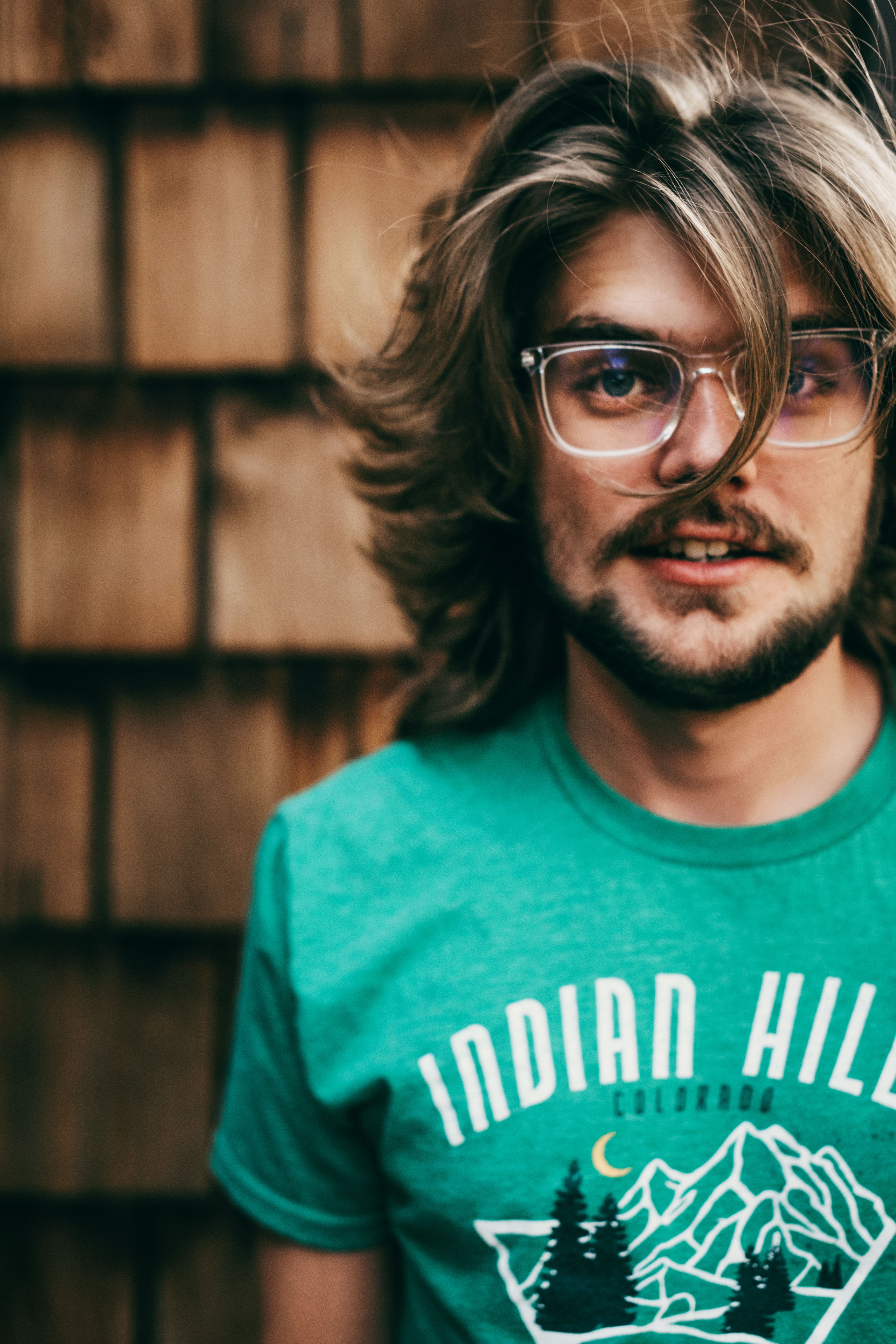 Portrait of a young man with long hair and glasses wearing a green shirt, standing outdoors.