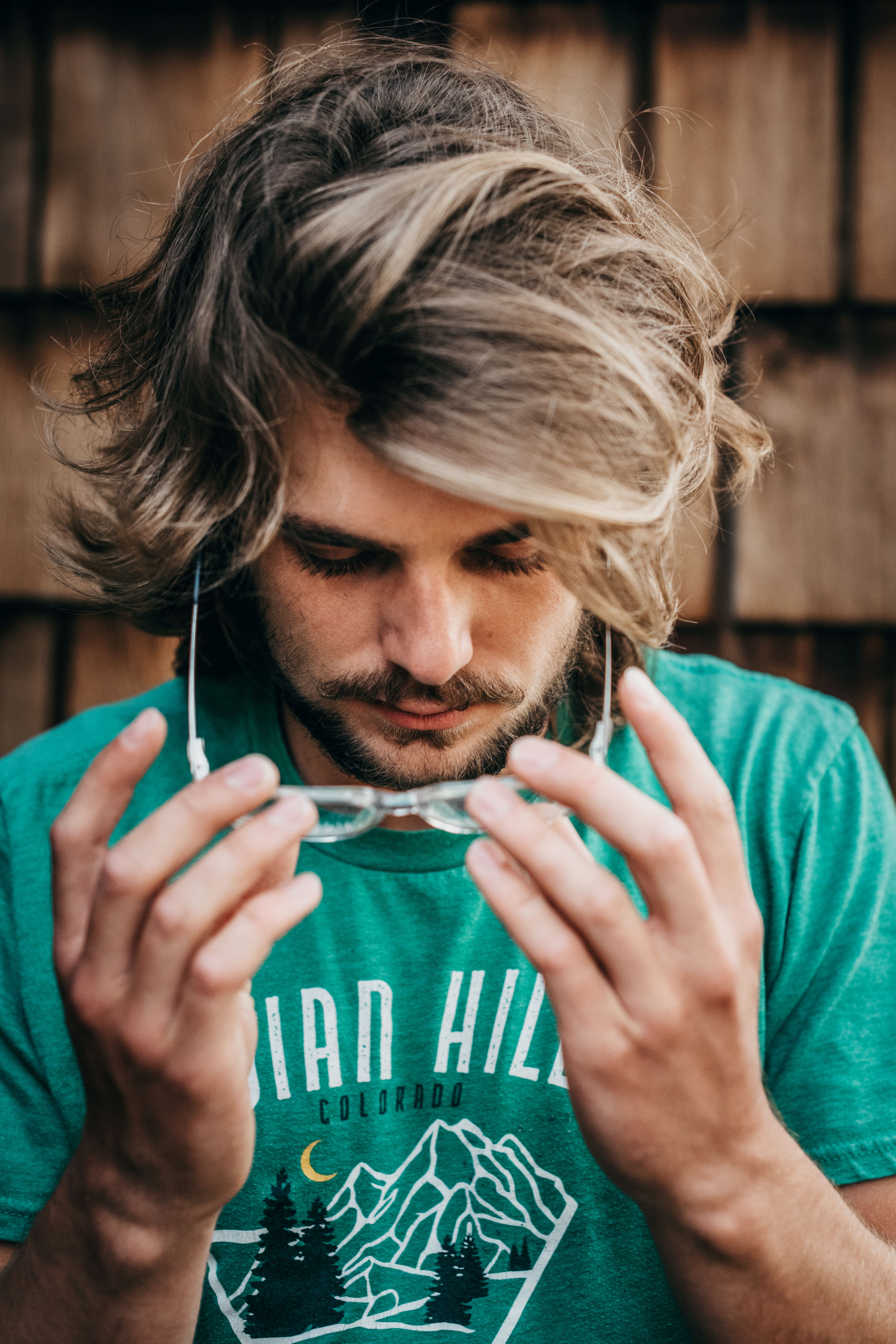 Young bearded man with long hair adjusting glasses in a casual setting.