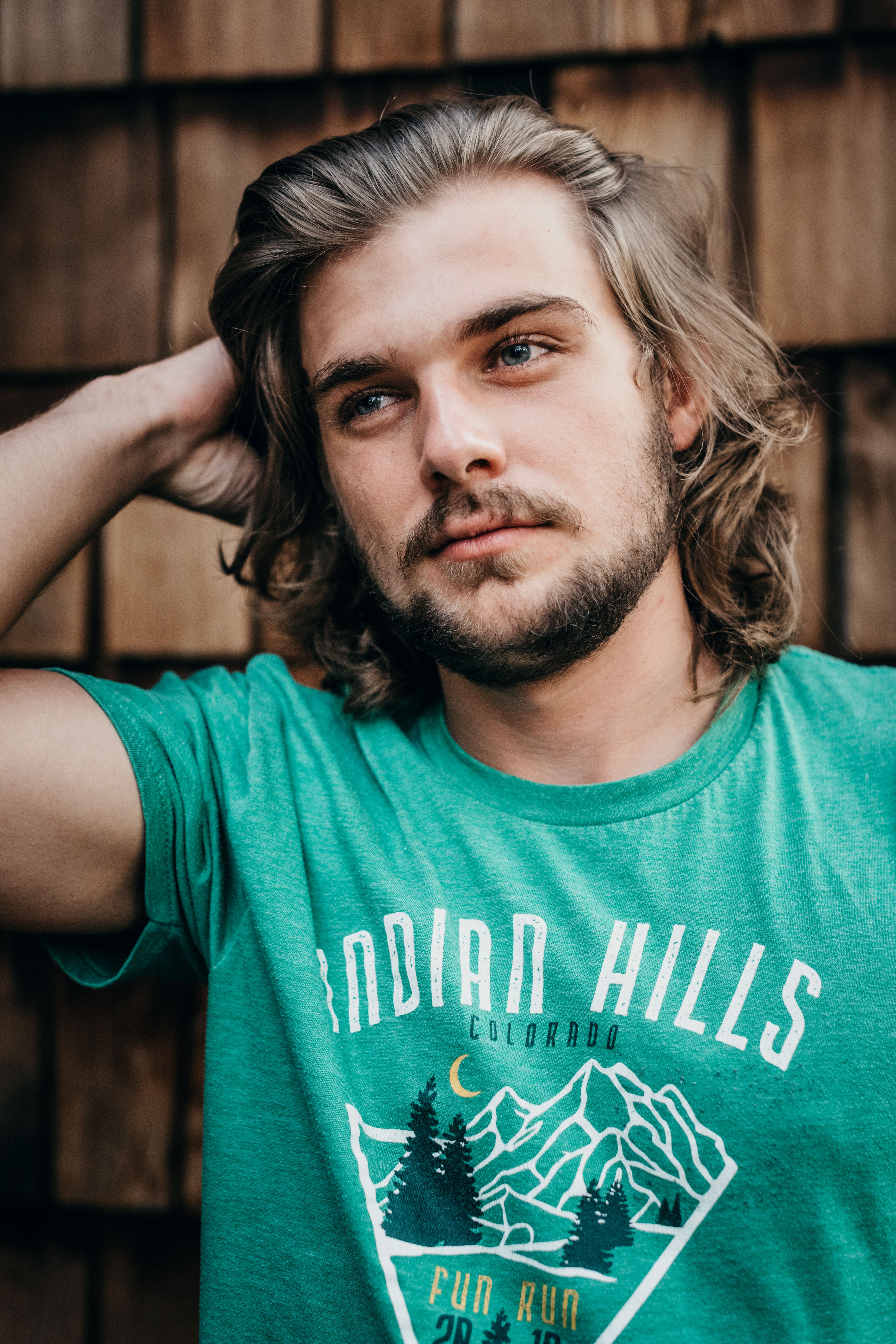 Portrait of a young man with long hair in a green shirt, set against a wooden background.