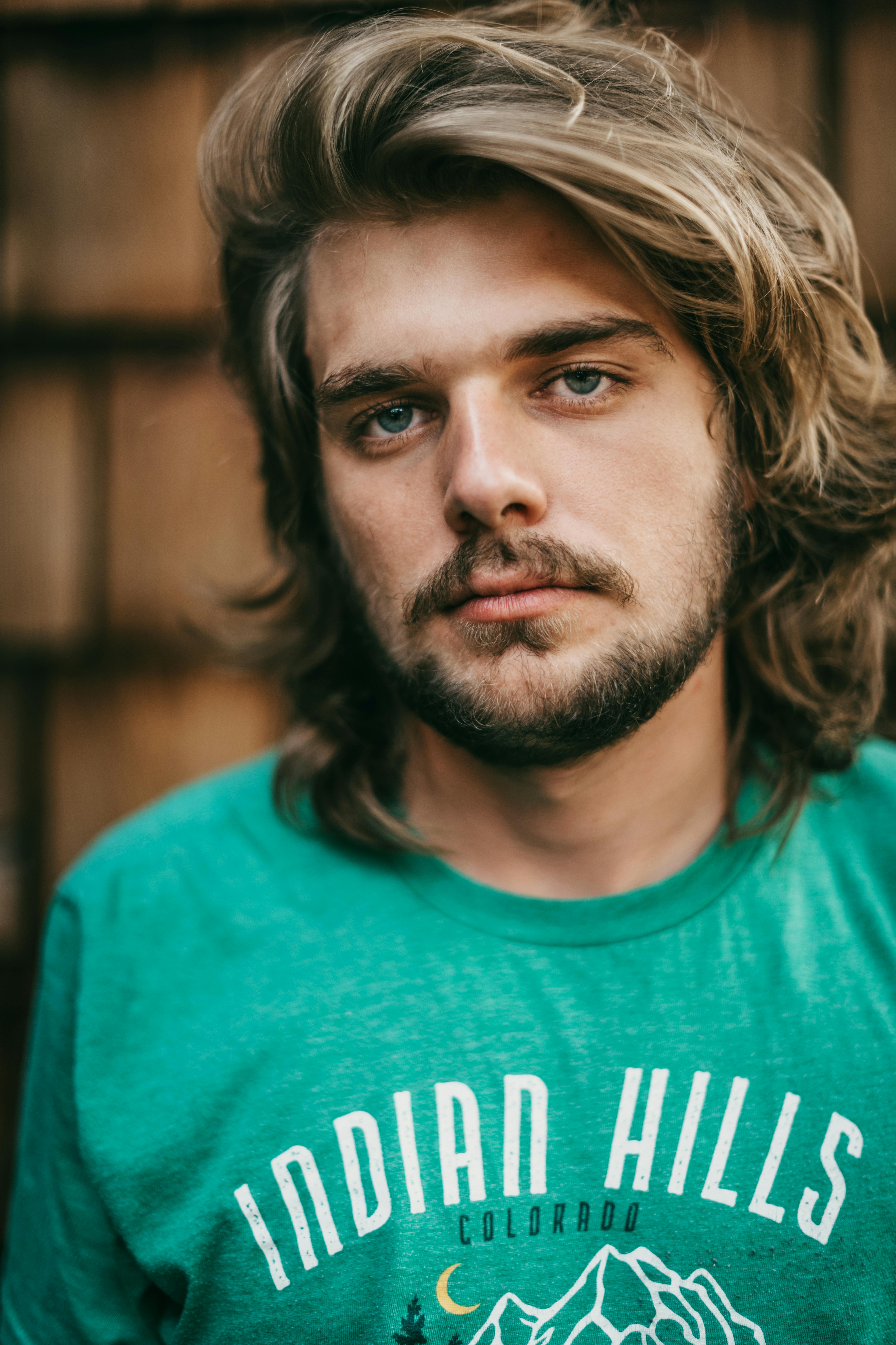 Close-up portrait of a bearded man in a green Indian Hills Colorado t-shirt with long blond hair.