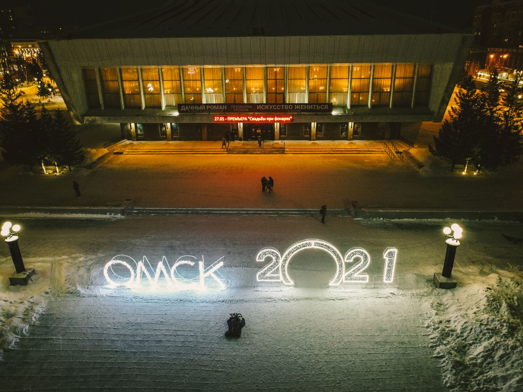 Aerial shot of illuminated Omsk theater during winter night with snow and festive lights.
