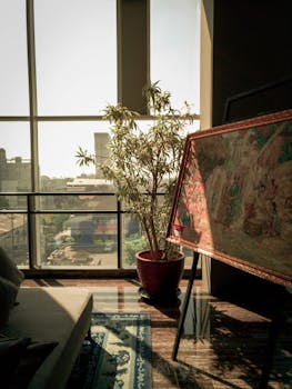 Cozy living room featuring large windows, a potted plant, and an artistic painting. Sunlight streams in.