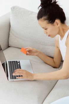 A woman shopping online using her credit card and laptop, lying on a sofa indoors.