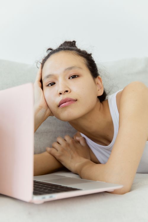 Free Portrait of a young woman lying on a couch using a laptop, wearing a white tank top. Stock Photo