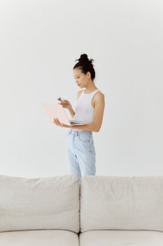Young woman holding a laptop in a minimalist room, dressed casually in jeans and a tank top.