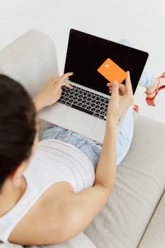 Woman holding orange credit card while shopping online on laptop from a sofa.