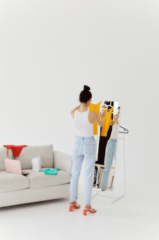 A woman in a white tank top considers a yellow blouse while online shopping at home.