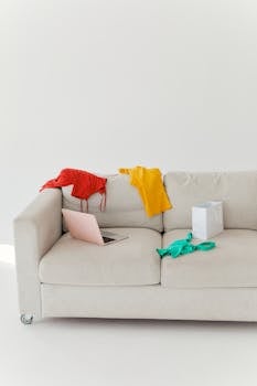 Cozy beige sofa with clothes, laptop, and shopping bag in bright minimalist room.