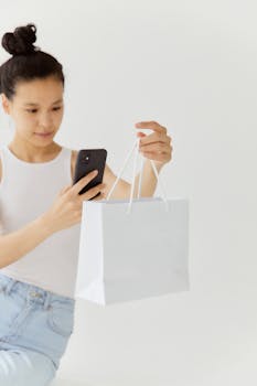 Asian woman photographing shopping bag with smartphone on white background.