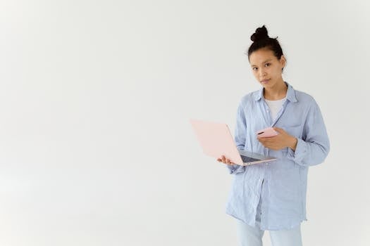 Young woman in casual attire holding a laptop and phone indoors against a minimalist background.
