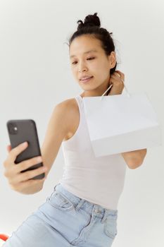 Asian woman in casual wear taking a selfie with a smartphone and white shopping bag indoors.